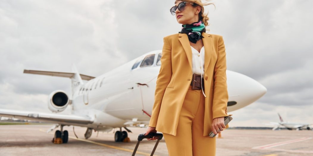 Passenger woman that is in yellow clothes, sunglasses and with luggage is outdoors near plane Passenger woman that is in yellow clothes, sunglasses and with luggage is outdoors near plane
