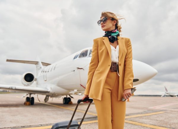 Passenger woman that is in yellow clothes, sunglasses and with luggage is outdoors near plane